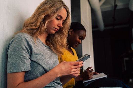 Blond businesswoman using smart phone while sitting with female colleague in office