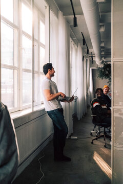 Young male programmer holding laptop talking with colleagues while standing near window in office