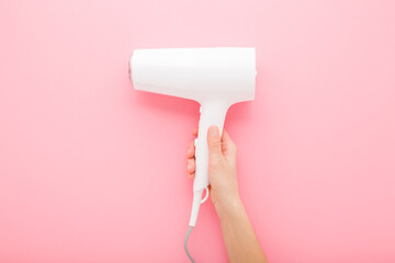 Young adult woman hand holding and showing new white hair dryer on light pink table background. Pastel color. Closeup. Top down view.