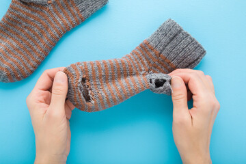 Young adult woman hands holding and showing ripped gray brown knitted sock on light blue table background. Pastel color. Closeup. Point of view shot. Flat lay. Top down view.