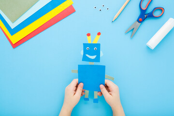 Little child boy hands holding and showing robot shape from colorful application paper on light blue table background. Pastel color. Paper craft. Closeup. Point of view shot. Flat lay. Top down view.