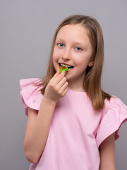 smiling young girl with fair skin and straight light brown hair is enjoying a gummy candy against a light gray studio background. She is wearing a soft pink shirt with ruffled sleeves and looking at t