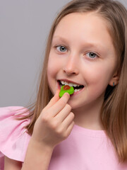 smiling young girl with fair skin and straight light brown hair is enjoying a gummy candy against a light gray studio background. She is wearing a soft pink shirt with ruffled sleeves and looking at t