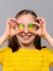 A cheerful young girl with light skin and brown hair in pigtails is posing against a light gray studio background. She is wearing a bright yellow knitted shirt and smiling broadly. The child playfully