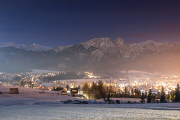 Night cityscape of Zakopane with the silhouette of Tatra Mountains. Illuminated mountain resort town in Poland under a dark evening sky.
