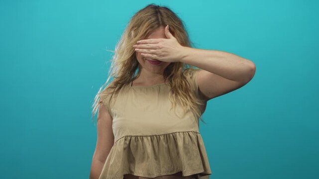 Woman blonde smiling, covering her eyes with hand and showing bare forearm in studio with turquoise backdrop and casual blouse; shyness.