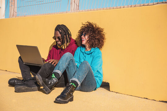 Cheerful multiethnic couple using laptop and having fun sitting on the ground near a yellow wall