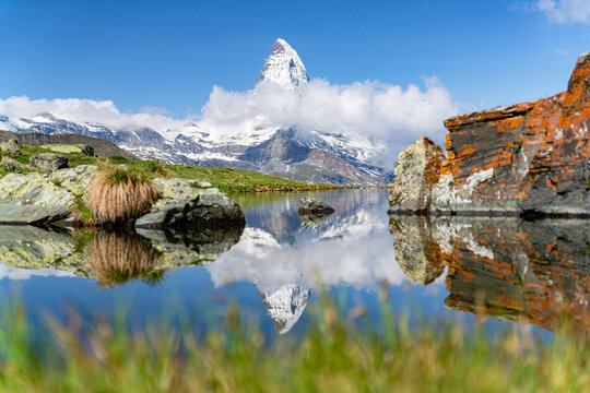 View of a serene Stellisee lake reflecting the snow-capped Matterhorn, framed by rugged, orange-tinged rocks and vibrant green meadows, Zermatt, Valais, Switzerland.