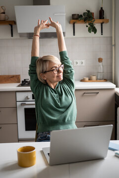 Satisfied woman warming up body after successfully finishing work tasks and projects on laptop. Cheerful female worker stretching arms muscles, taking break from work on computer at kitchen home. 