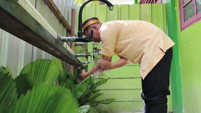 
Asian Muslim man wearing a yellow shirt, black pants, and traditional cap performing ablution by washing his arms, representing Islamic ritual purification before prayer