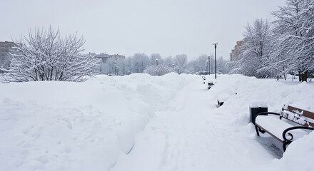 Urban pedestrian zone blocked by accumulated snow, benches and pathways barely visible under deep layers. Winter scene displays vast landscape, urban infrastructure reflecting accumulated snow.