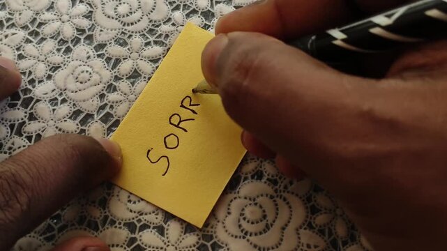 Indian or Asian person hand with pen writing sorry apologize message or word on blank sticky note paper on a desk background. Sorry handwritten calligraphy text card concept.