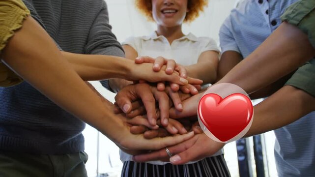 Red heart icon over mid section of diverse office colleagues staking their hands together at office
