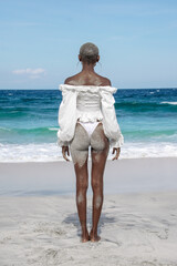 Back view of a woman with short textured hair and hoop earrings standing on a tropical beach, looking at the ocean. Minimalist summer scene symbolizing freedom, calm, travel, self-reflection, and conn