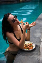 Young woman wearing sunglasses eating shrimp in a swimming pool, summer lifestyle concept