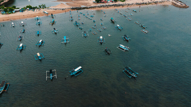 Drone aerial view of traditional Balinese outrigger fishing boats anchored in shallow sea near the shoreline at Mertasari Beach, Sanur, Bali, Indonesia.