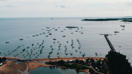 Drone aerial view of traditional Balinese outrigger fishing boats anchored off Mertasari Beach with a long pier extending into the sea in Sanur, Bali, Indonesia.