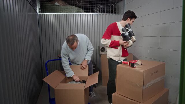 Father and son organizing seasonal clothing and sports equipment in self storage unit, unpacking winter jacket from garment bag and talking while standing among boxes