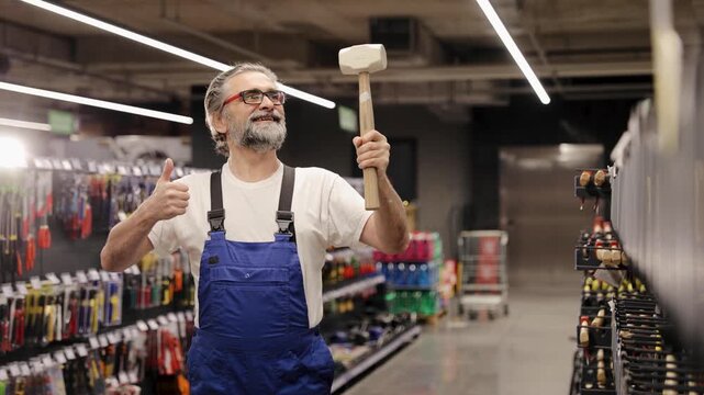 Experienced handyman in overalls wearing glasses and a beard, standing confidently with a hammer in a retail hardware environment