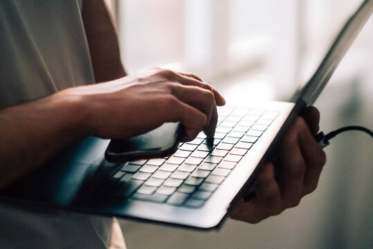Hand of businessman holding smart phone while pressing laptop key