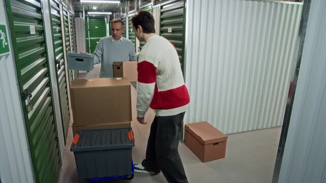 Middle age man and his teenage son loading cardboard boxes and plastic container onto blue cart, then pushing it down hallway in indoor storage facility