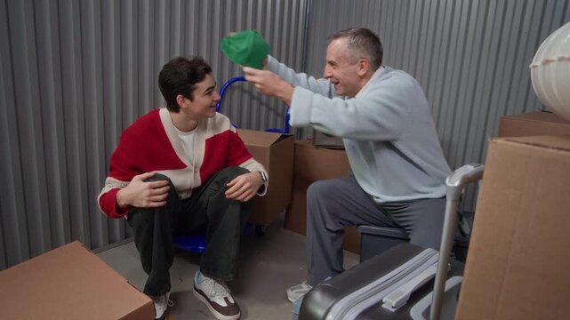 Cheerful middle aged father and young son sitting together in storage unit surrounded by cardboard boxes, discussing green cap and trying it on