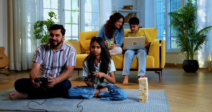 Indian father daughter playing video game and mother son sitting on sofa behind as family enjoys fun time together indoors in modern lavish home living room lifestyle scene