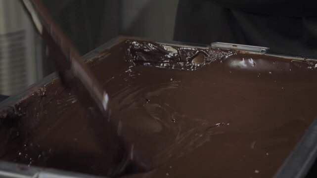 Close-up of thick melted dark chocolate pouring into metal tray creating glossy swirls in factory.