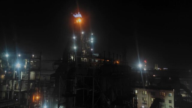 Static night view of an industrial refinery with illuminated structures and a flare burning atop a tower, casting light over pipes, platforms, and surrounding facilities.