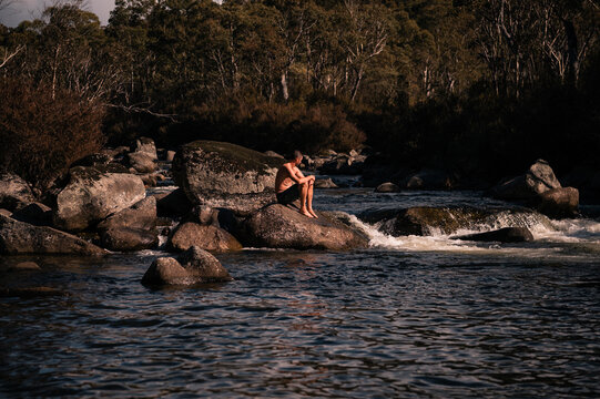 A man sits on a rock in the middle of a river on a winter day