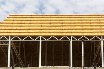 Fototapeta premium Building under construction featuring a geometric roof structure. Yellow OSB panels and wooden beams sit on a grey metal frame. Industrial, architectural background against a cloudy blue sky.