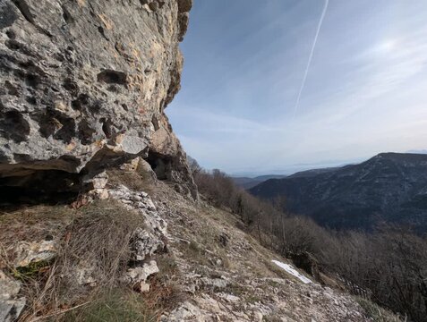 Paesaggio montano con ripida parete rocciosa, terreno arido e vegetazione rada, con vista su colline e montagne lontane sotto un cielo limpido attraversato da una scia aerea