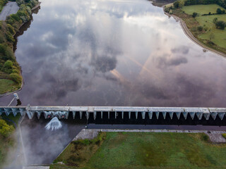 High-angle aerial view of Val Joly dam and its reservoir, with clouds reflected on the calm water, showcasing engineering, natural scenery, and tranquil landscapes in northern France.

