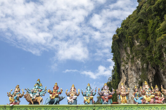 View of vibrant Hindu deities adorn the temple entrance against the dramatic backdrop of towering limestone cliffs and a bright blue sky, Batu Caves, Selangor, Malaysia.