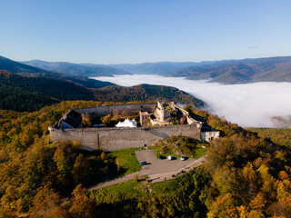 Medium aerial view of Ch&acirc;teau du Hohlandsbourg perched on a hill, overlooking a valley filled with low white clouds under a clear blue sky in the Alsace region of France.

