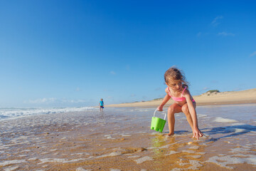 Happy blond girl collecting seashells on beach with green bucket