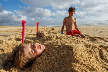 Boy and girl playing on beach, buried in sand by teen sibling