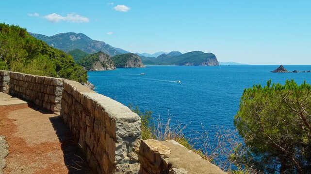 Scenic coastal view of the Adriatic Sea from a stone promenade in Petrovac, Montenegro. Stunning summer landscape featuring turquoise water, rocky cliffs, pine trees, and a clear blue sky
