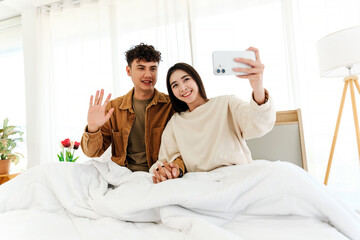 Obraz premium A cheerful couple takes a selfie in bed, smiling and waving with warmth and joy. Surrounded by cozy light, the moment captures youthful love, connection, and relaxed togetherness.
