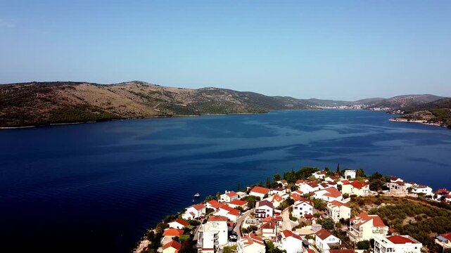 Wide aerial pullback of coastal village and harbor with red roofs beside deep blue Adriatic water, Seget Vranjica Croatia