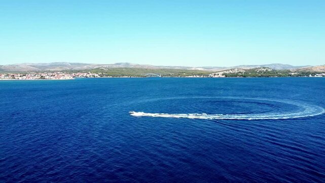 Circular wake pattern on calm sea created by turning boat pulling tube riders, aerial pan