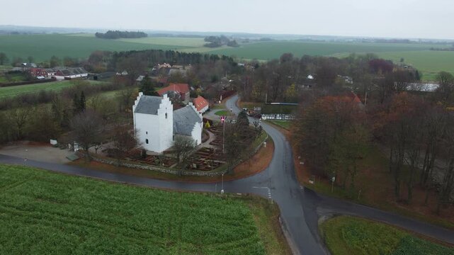 Picturesque white Vejlby church standing beside a country road and surrounded by a graveyard and lush green fields in the rural landscape of Grenaa, Denmark on an overcast day