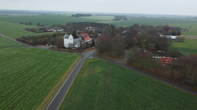 High angle view of a vast green landscape with a small village, a white car drives along a country road passing a characteristic medieval church, revealing the serene and scenic rural area