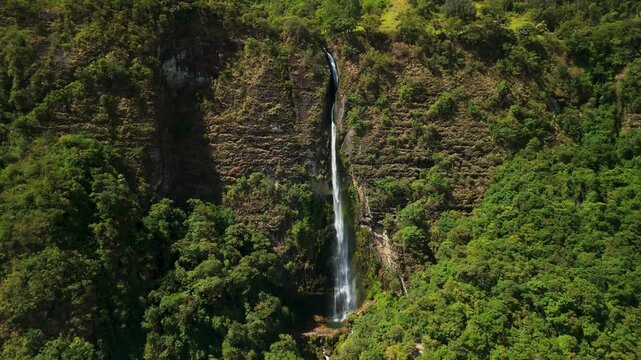 Cinematic drone shot retreats from cascading waterfall to reveal massive vertical cliff face and dense tropical greenery surrounding deep rocky canyon in Azuay province