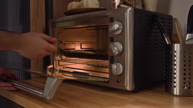 Zoom in view of unrecognisable young man placing baking sheet with homemade pizza into preheated electric oven