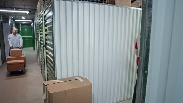 Young tenant stacking cardboard boxes while unloading his storage unit, middle aged man pushing cart through hallway and passing by him