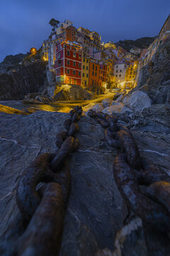 View of ancient chains lead the eye to a cascade of vibrant, stacked houses clinging to the cliffside at dusk, glowing warmly against the twilight, Riomaggiore, Liguria, Italy.
