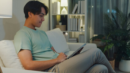 Young Asia male with casual cloth sit on sofa finger scroll digital tablet watch online movie in living room cozy house at night. E-book reading, Shopping online, Lifestyle leisure at home concept.
