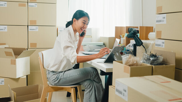 Youth attractive Asia female entrepreneur small business start up call smartphone confirm order with customer confirm check stock inventory product ready for delivery shipment at online retail shop.