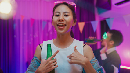 Diverse multi ethnic young adult Asian lady wear futuristic neon glowing glasses smile and drink alcohol beer fun enjoy hangout party in living room night indoor home. Disco lights illuminating house.